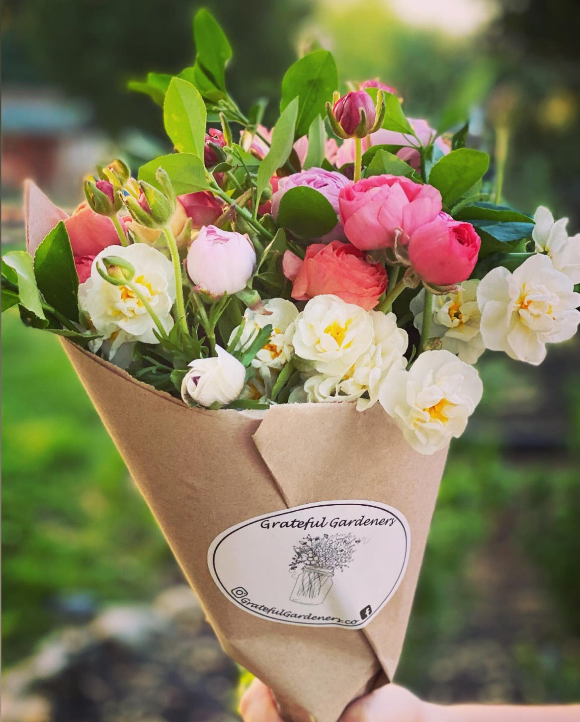 Bouquet of flowers wrapped in brown paper with a visible brand label, set against a blurred natural background.